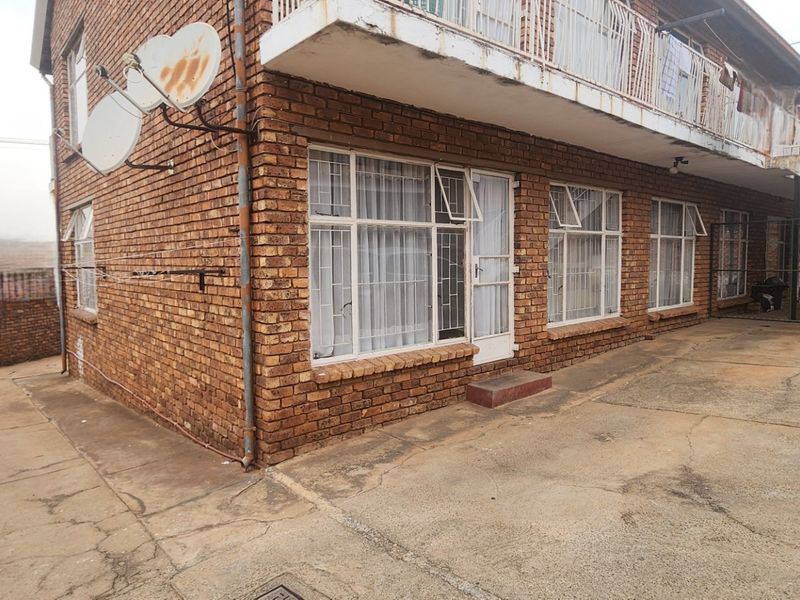 Exterior view of a multi-story apartment building featuring face brick construction, a concrete balcony with metal railings, and paved ground level access.