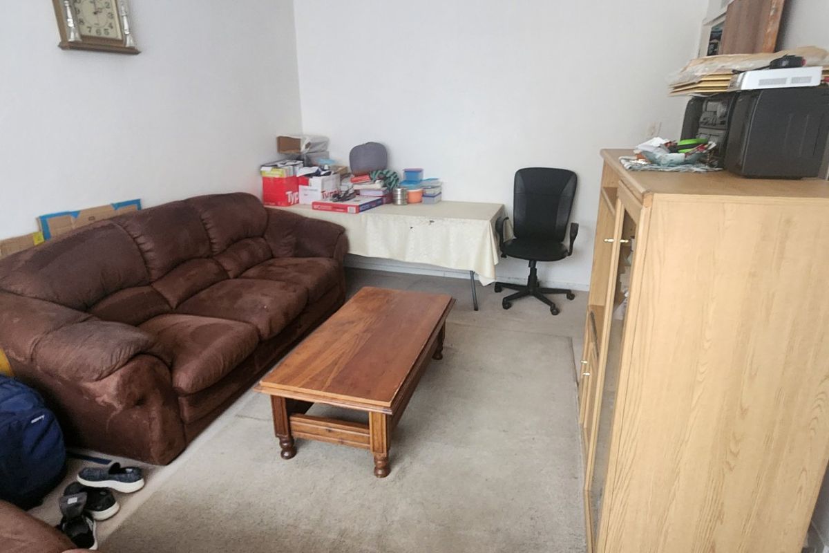 A living room space with plain walls and carpeted flooring, currently utilized as a multi-purpose area.