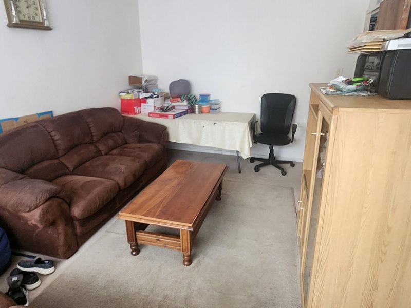 A living room space with plain walls and carpeted flooring, currently utilized as a multi-purpose area.