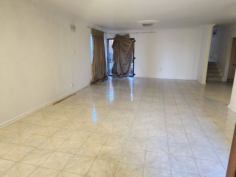 A spacious, empty living area featuring neutral ceramic floor tiles, a ceiling rose, and access to an outdoor area via a security gate.