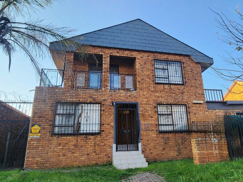 Two-story face brick apartment building featuring a tiled roof, security bars on windows, and an upper-level balcony with glass balustrades.