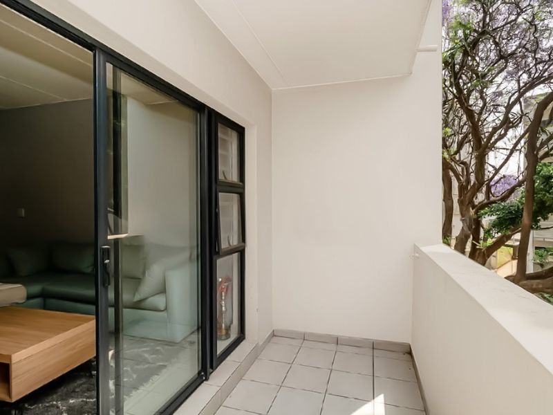A clean, covered balcony featuring tiled flooring and a solid balustrade, accessible via large sliding glass doors from the living area.