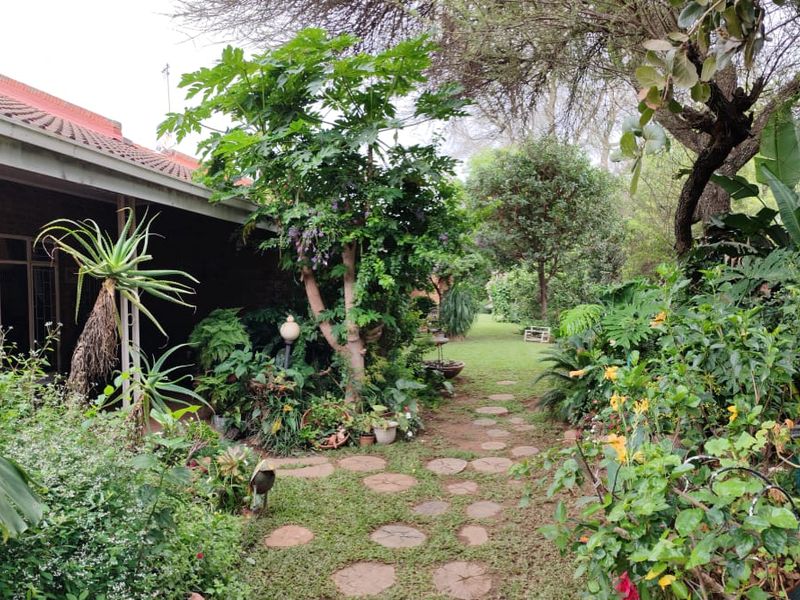 A lush, established garden featuring a stone paver pathway leading alongside a brick house with a covered patio and mature trees.