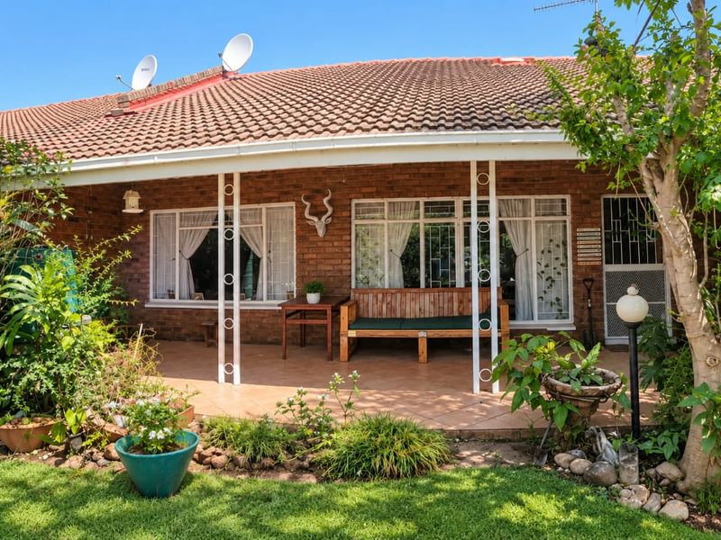 A covered patio area featuring tiled flooring, brick exterior walls, and metal support columns, overlooking a landscaped garden.