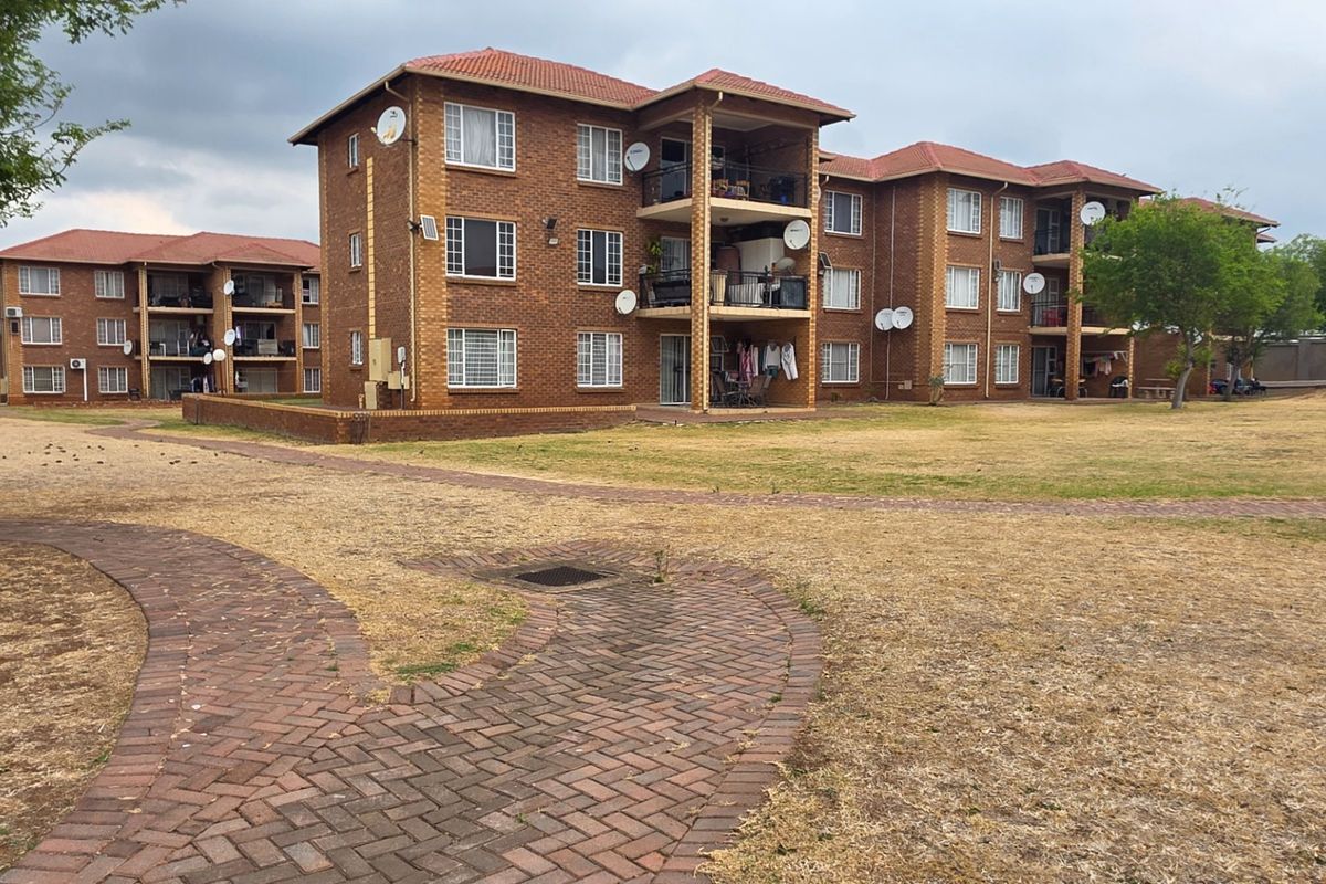 Exterior view of a multi-story face brick apartment complex featuring tiled roofs, private balconies, and a paved walkway through a communal garden area.
