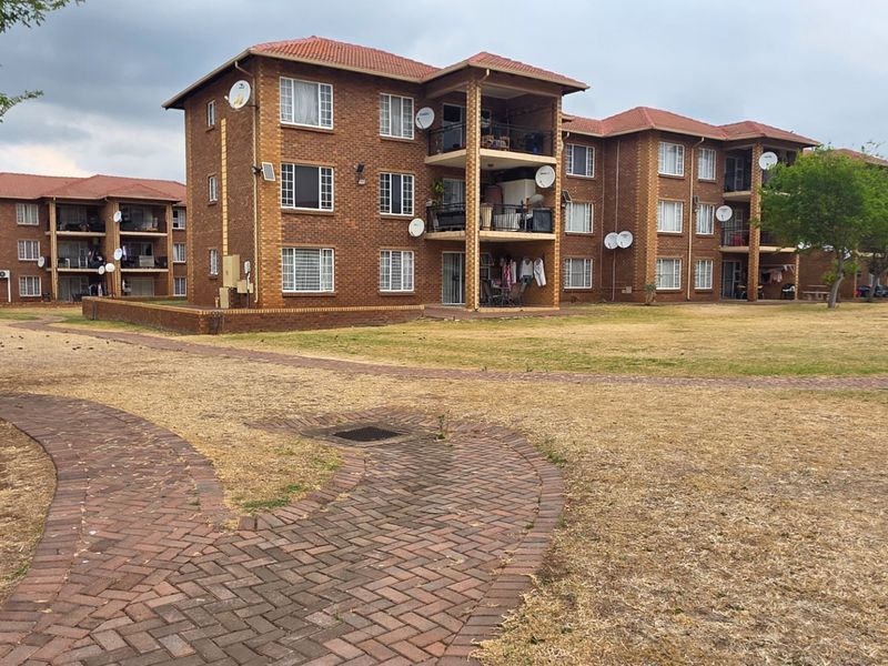 Exterior view of a multi-story face brick apartment complex featuring tiled roofs, private balconies, and a paved walkway through a communal garden area.