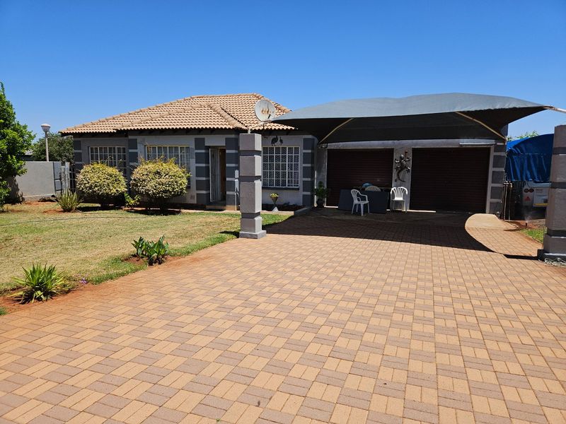 Front view of a single-story house featuring a paved driveway, a large shade cloth carport, and a well-maintained garden.