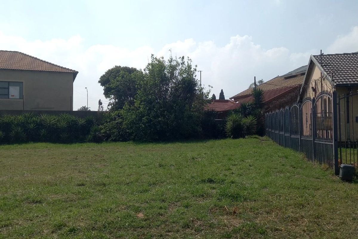 A vacant residential plot featuring a grass lawn and metal perimeter fencing, situated between existing residential properties.