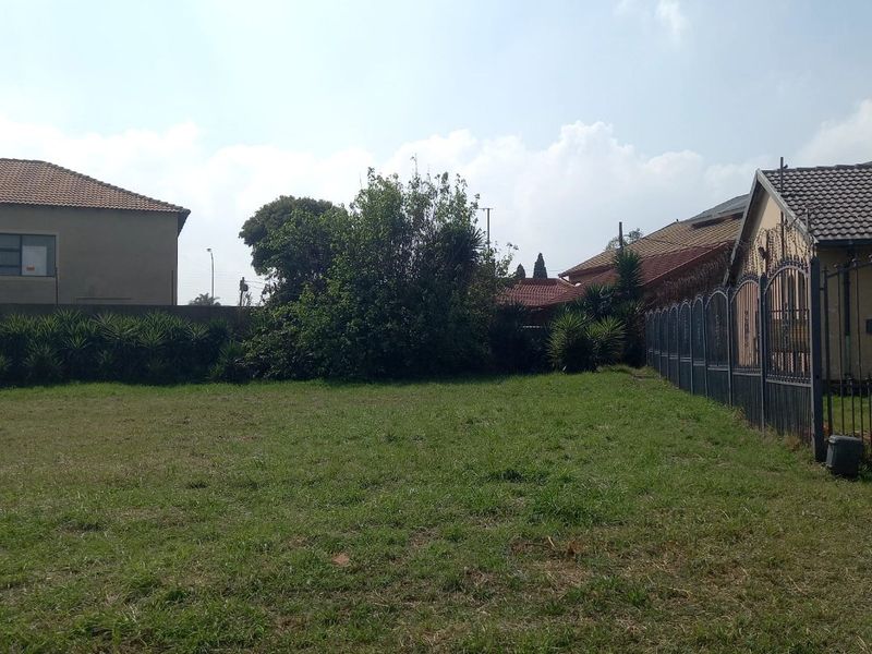 A vacant residential plot featuring a grass lawn and metal perimeter fencing, situated between existing residential properties.