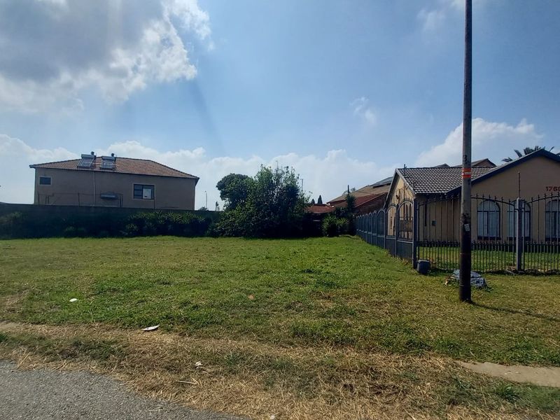 A vacant residential plot of land featuring grass coverage, enclosed by perimeter fencing, and situated adjacent to existing residential properties.