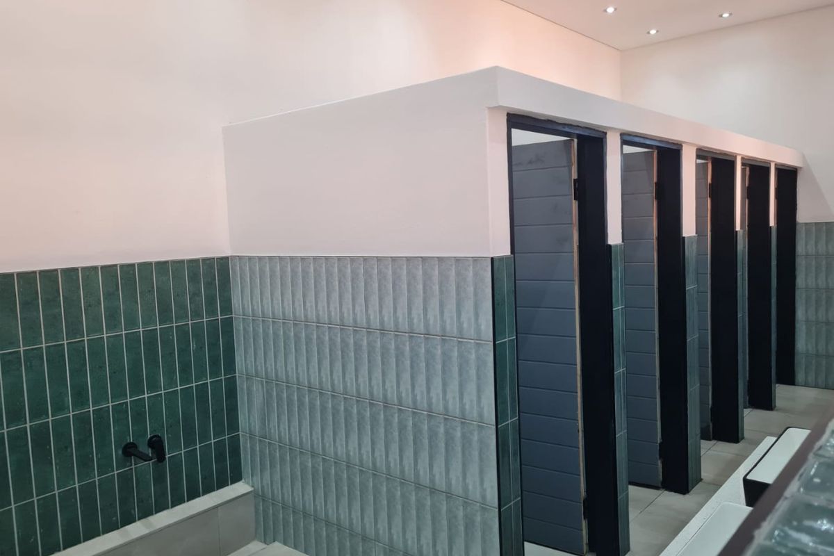 Modern commercial bathroom facility featuring partitioned toilet cubicles, stylish green vertical tiling, and recessed ceiling lighting.