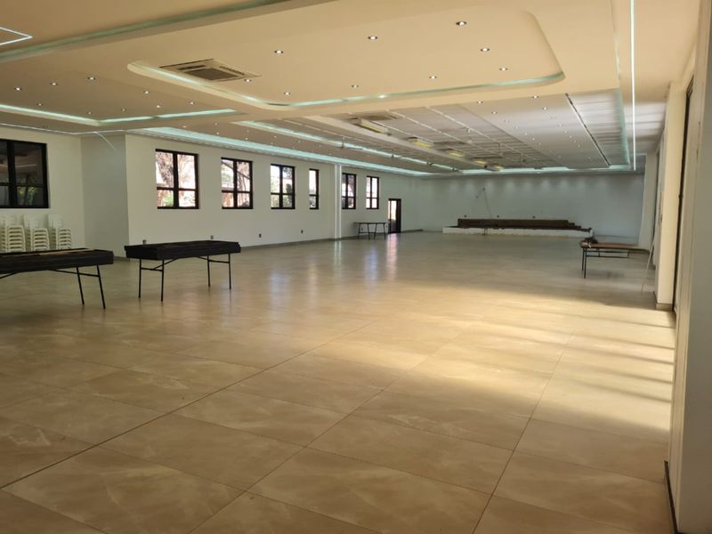 Spacious open-plan commercial hall featuring large windows, recessed ceiling lighting, tiled flooring, and a raised stage area suitable for events or conferences.