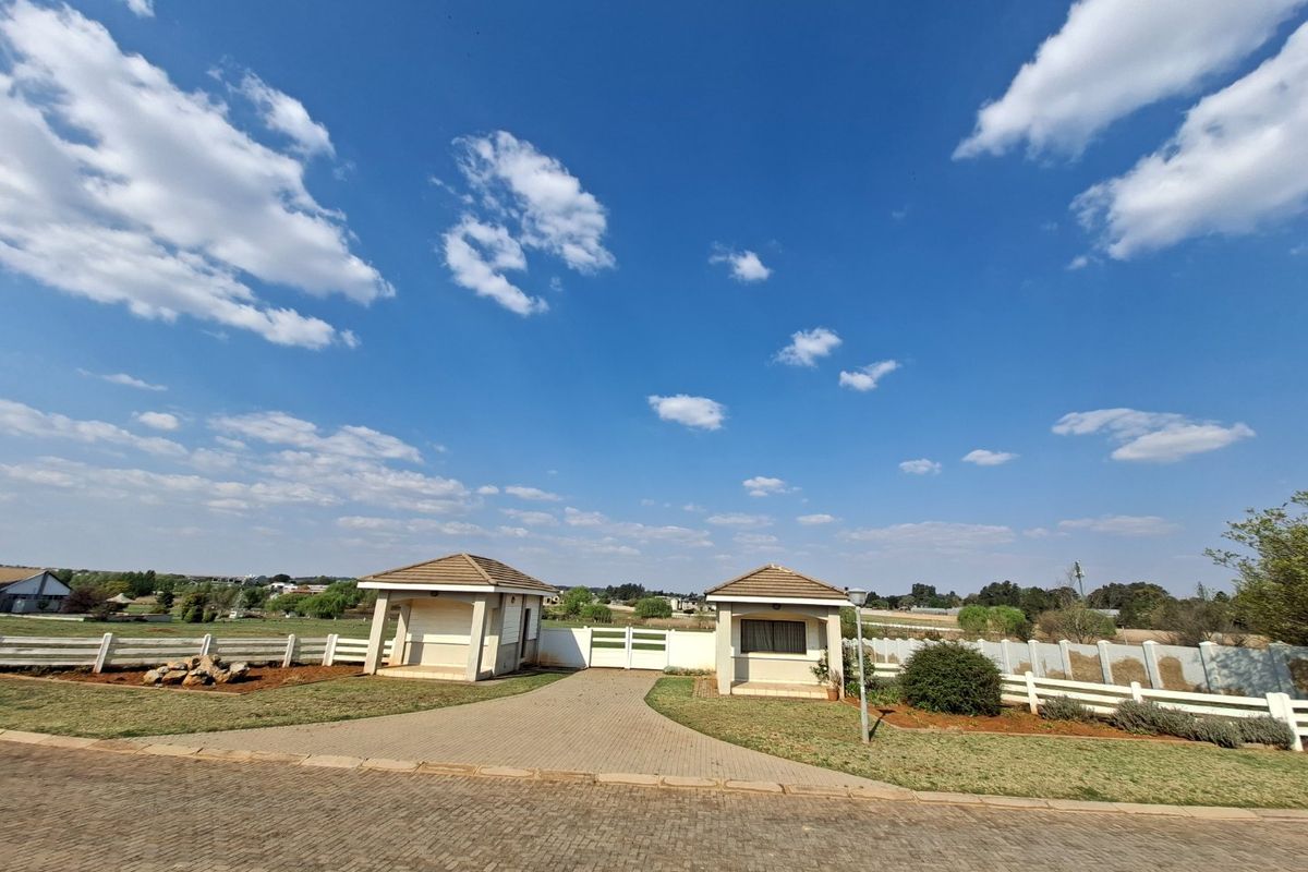 A well-maintained residential estate entrance featuring a paved driveway, security gatehouse, and perimeter fencing set against a clear blue sky.