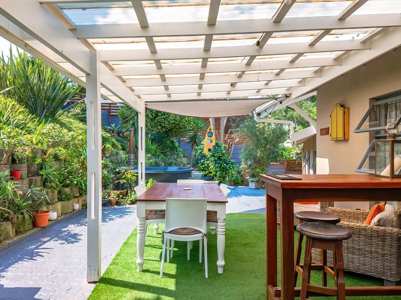 A covered outdoor patio area featuring a pergola with translucent roofing, artificial turf, and a tiered garden retaining wall.