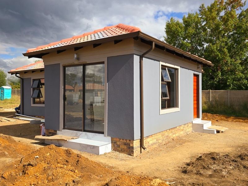 A modern exterior view of a newly constructed house featuring a tiled roof, grey rendered walls, and sliding glass doors, currently awaiting landscaping.