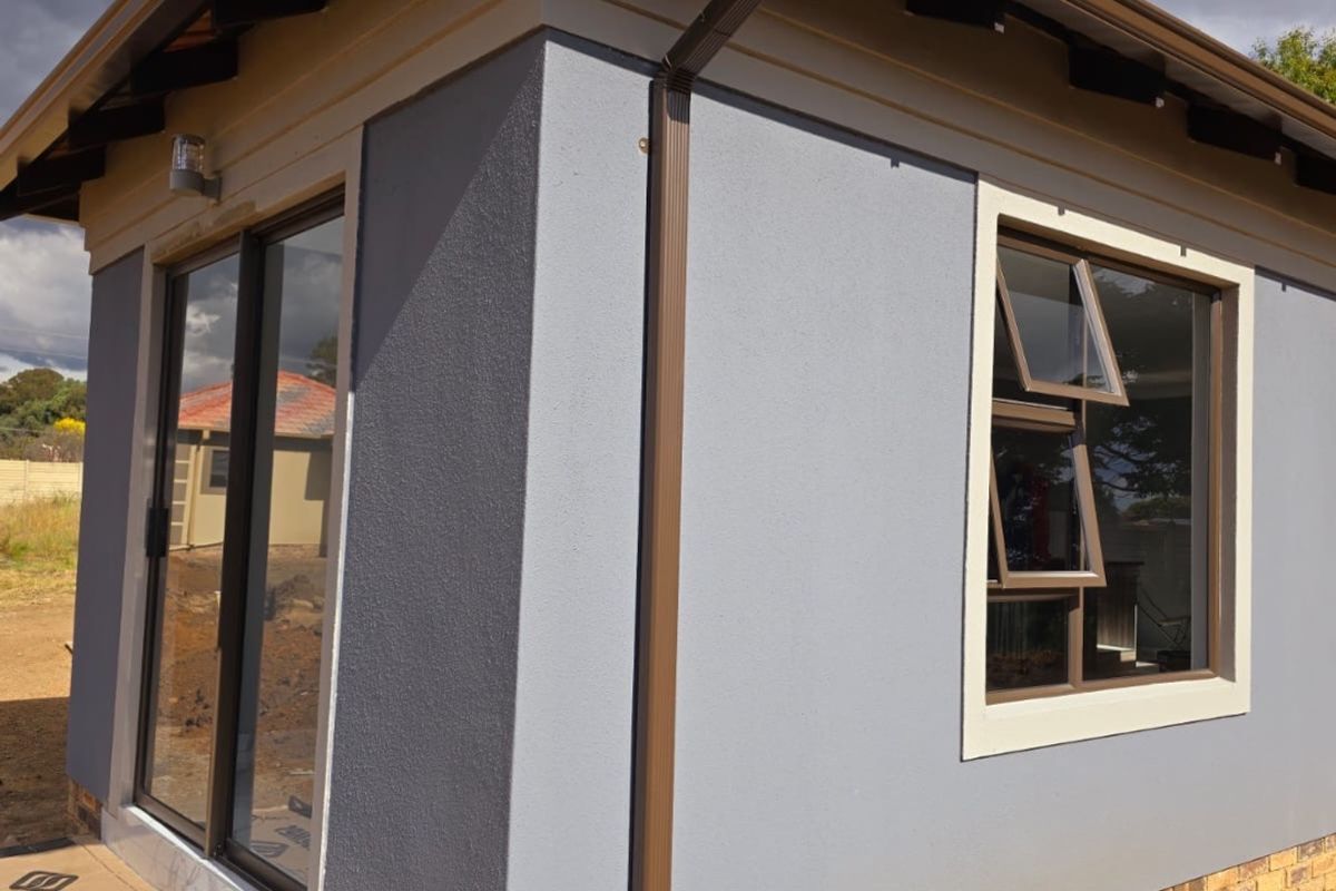 A modern exterior view of a newly constructed building featuring grey textured walls, a brick base, sliding glass doors, and a tiled roof.