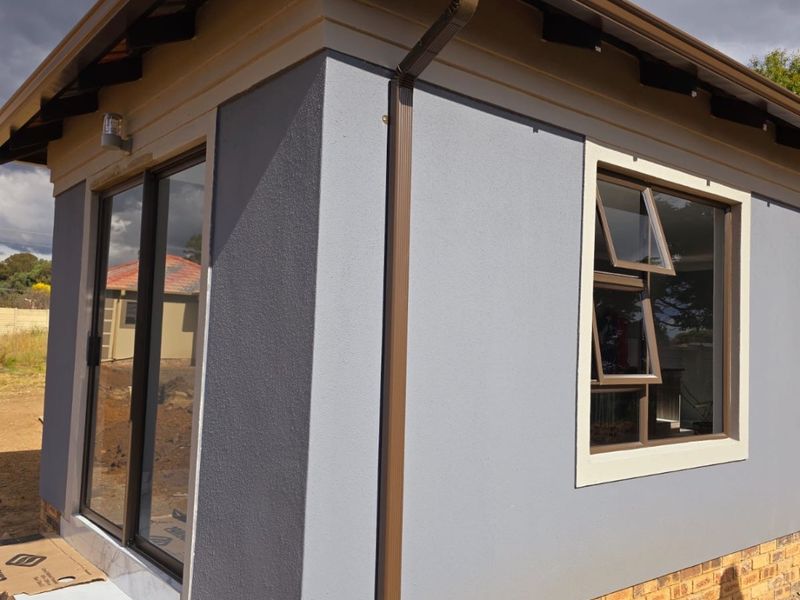 A modern exterior view of a newly constructed building featuring grey textured walls, a brick base, sliding glass doors, and a tiled roof.