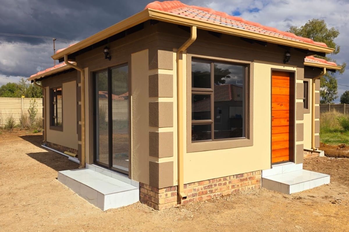 A modern detached structure featuring a tiled roof, aluminium window frames, sliding glass doors, and a wooden entrance door, set on an unfinished plot.
