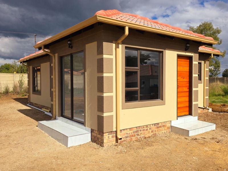 A modern detached structure featuring a tiled roof, aluminium window frames, sliding glass doors, and a wooden entrance door, set on an unfinished plot.