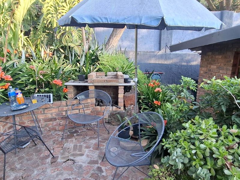 A cozy outdoor patio area featuring a built-in brick braai, established garden landscaping, and brick paving, perfect for relaxed outdoor entertaining.