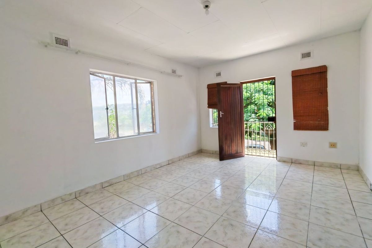 A bright, empty living area featuring tiled flooring, natural light through aluminium windows, and direct access to an outdoor space via a security gate.