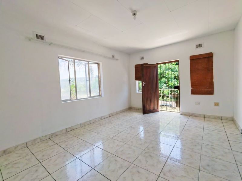 A bright, empty living area featuring tiled flooring, natural light through aluminium windows, and direct access to an outdoor space via a security gate.