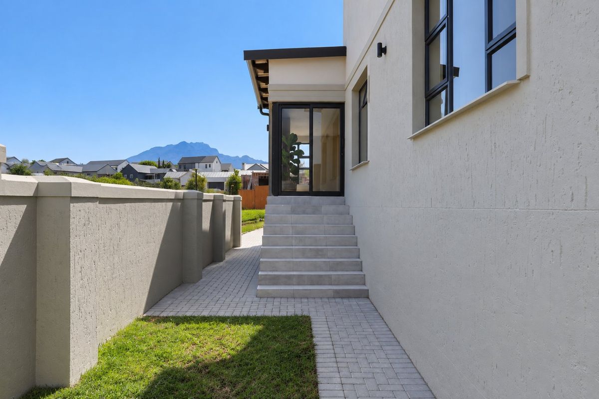A well-maintained rear exterior view featuring a paved walkway, concrete steps leading to a sliding glass door, and a solid boundary wall.