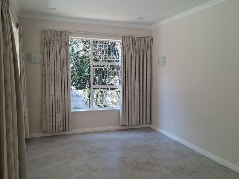 A bright, empty living room featuring neutral tiled flooring, crown molding, recessed lighting, and large windows with security bars.