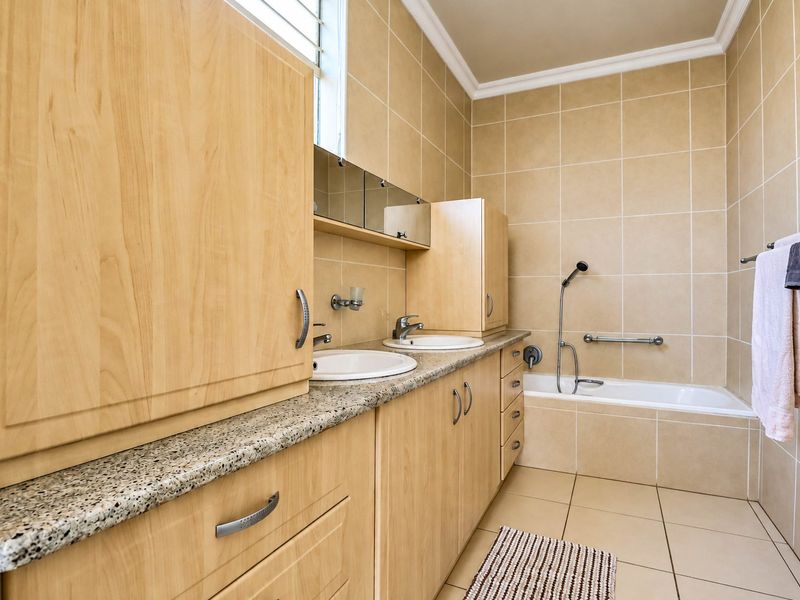 A functional bathroom featuring a double vanity with granite countertops, built-in light wood cabinetry, and a tiled bathtub area with a handheld shower fixture.