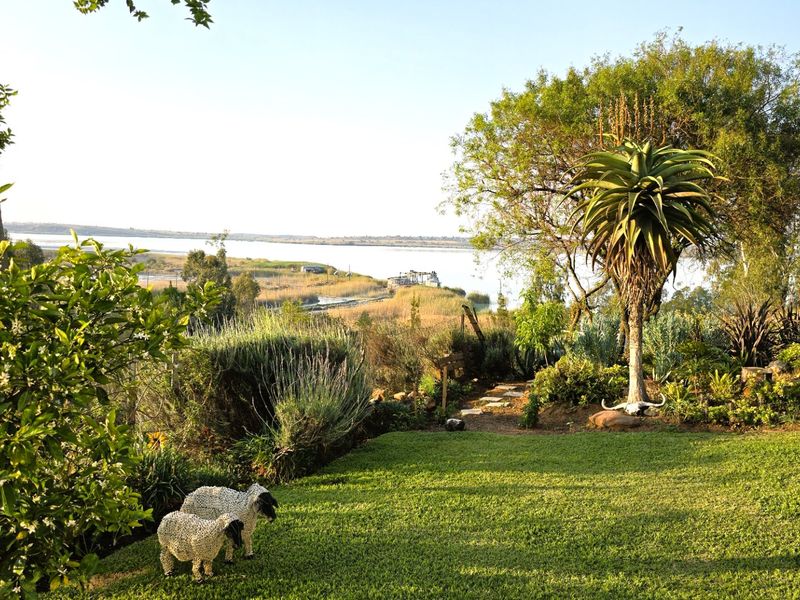 A scenic view of a landscaped garden featuring mature trees and aloe plants, overlooking a tranquil lake and natural landscape.
