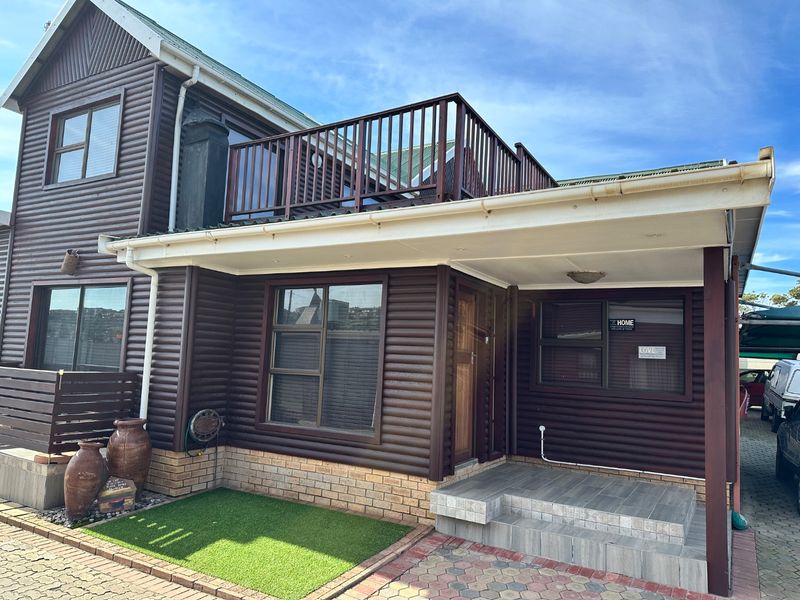 Front elevation of a two-story house featuring timber cladding, a brick base, and an upper-level balcony with a paved entrance area.
