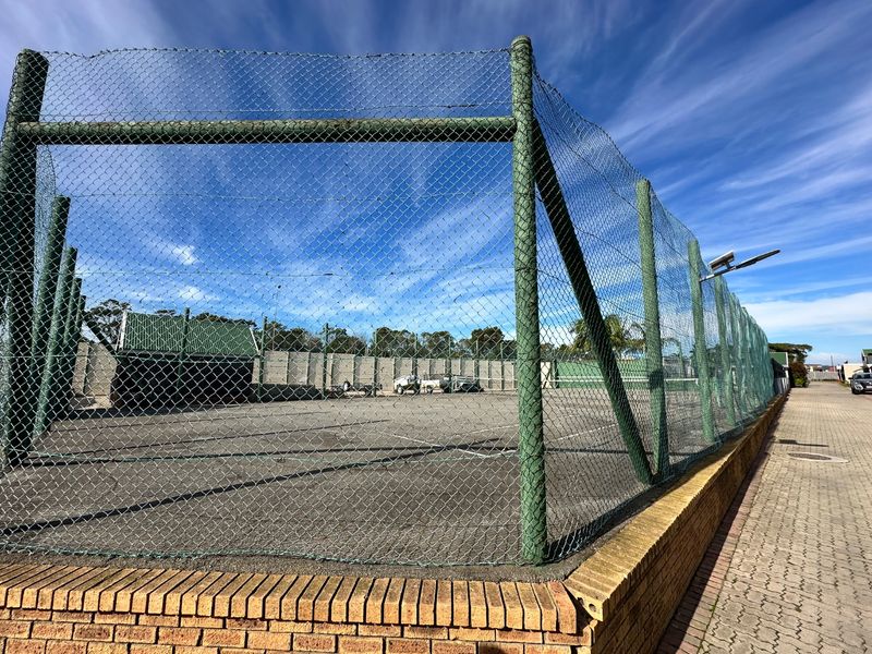 A fenced outdoor sports court or paved recreational area featuring a brick retaining wall and security lighting.
