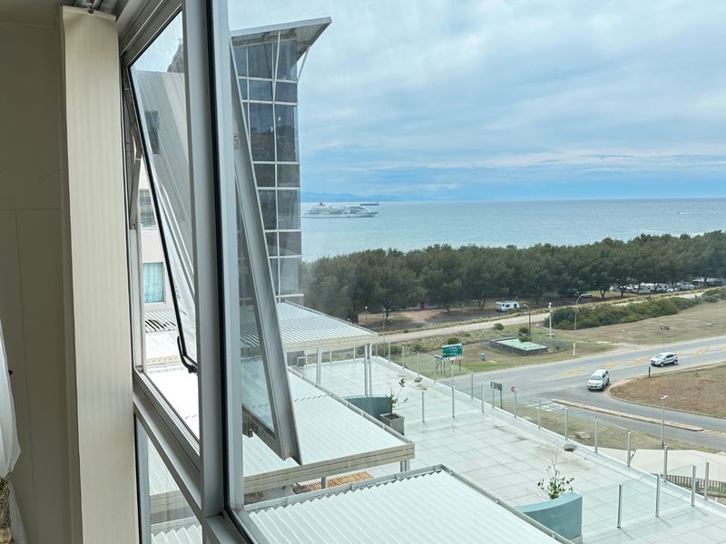 Elevated view from an apartment window overlooking the coastline, nearby road, and ocean horizon.
