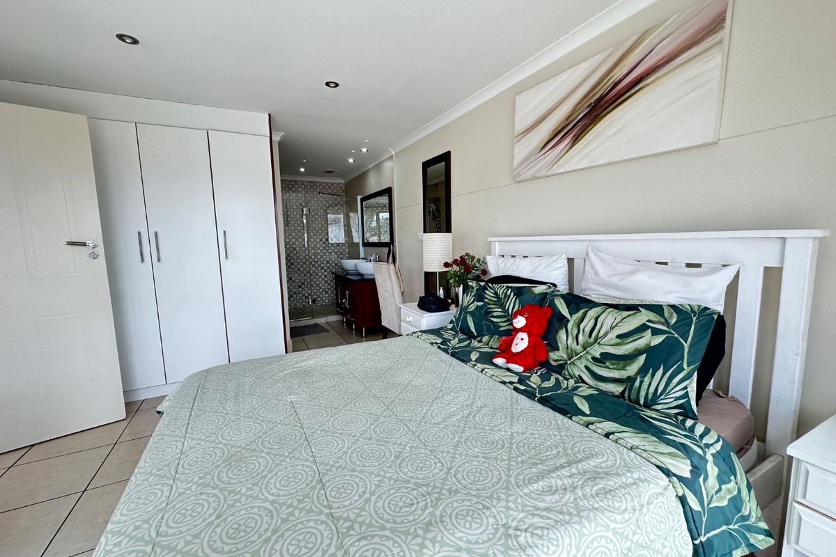 A well-maintained bedroom featuring built-in cupboards, tiled flooring, recessed lighting, and an open-plan en-suite bathroom with a double vanity and glass shower.