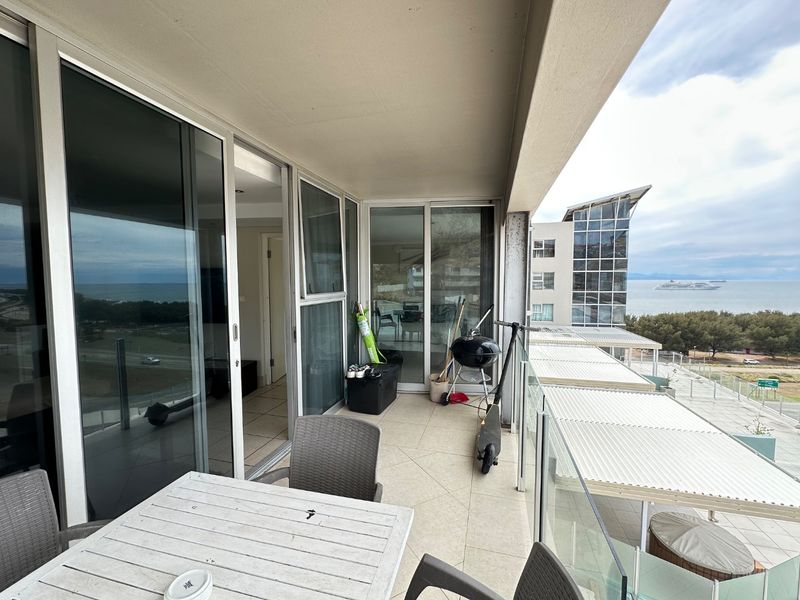 A spacious covered balcony featuring tiled flooring, a glass balustrade, and sliding door access, offering expansive coastal and neighborhood views.