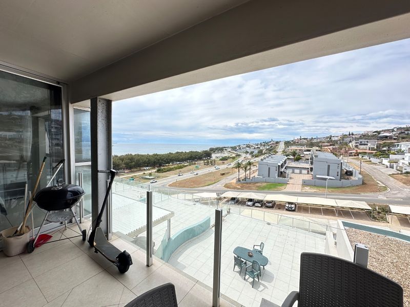 A spacious covered balcony featuring a glass balustrade and tiled flooring, offering expansive coastal and neighbourhood views.