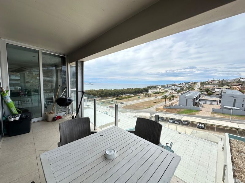 Spacious covered balcony featuring a glass balustrade and tiled flooring, offering expansive coastal and neighbourhood views through large sliding glass doors.
