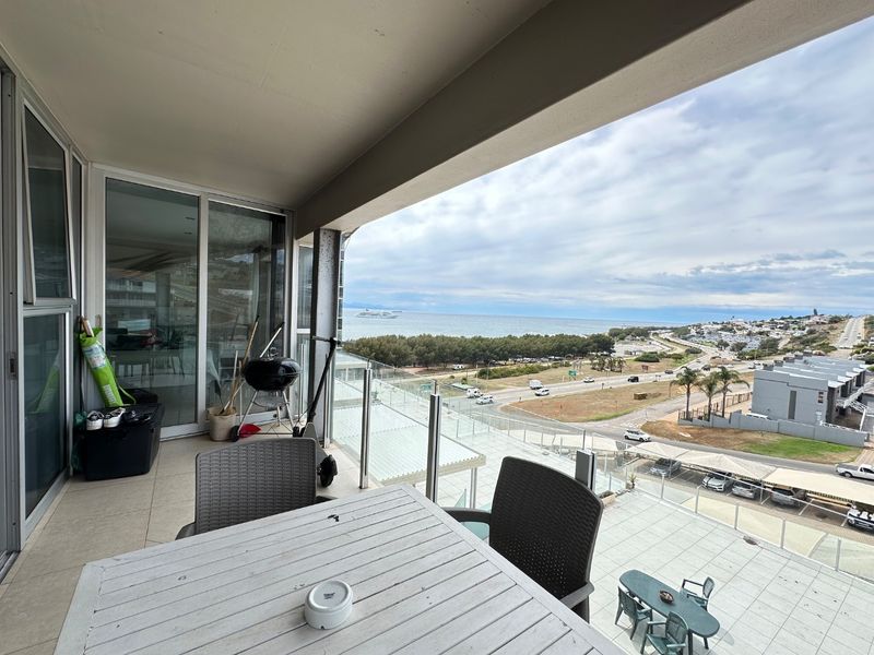 Spacious covered balcony featuring a glass balustrade and expansive coastal and road views, accessible via large sliding glass doors.