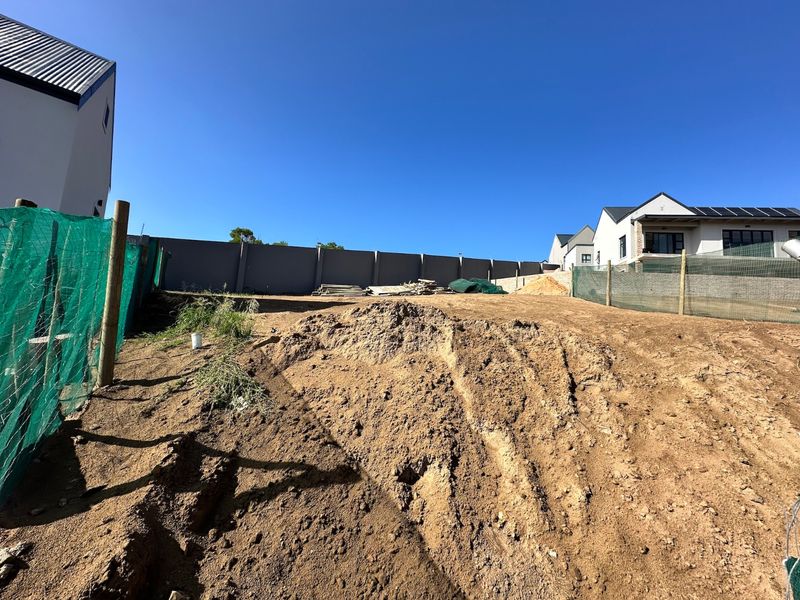 A vacant residential plot featuring a sloped terrain, perimeter boundary wall, and temporary construction fencing, ready for development.