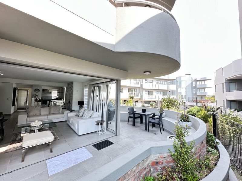 An open-plan living area seamlessly connecting to a spacious curved balcony, featuring modern tiled flooring and large sliding glass doors for indoor-outdoor flow.