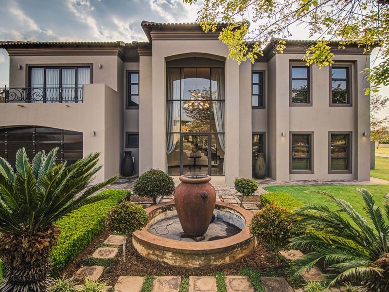 Impressive double-storey residence featuring a grand glass entrance, manicured garden with a central stone fountain, and a paved driveway.