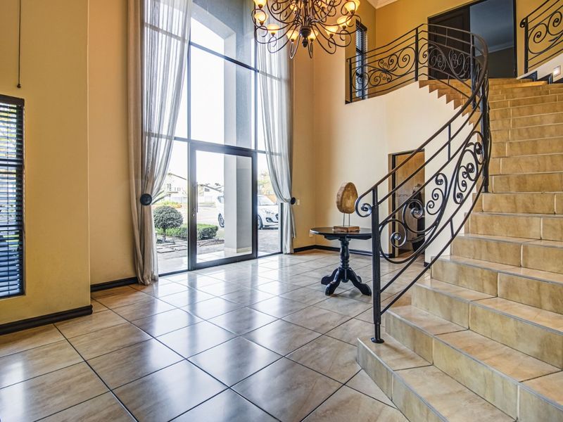Grand double-volume entrance hall featuring a sweeping tiled staircase with ornate wrought iron balustrades, large floor-to-ceiling windows, and elegant chandelier lighting.