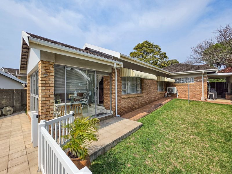 Rear view of a face brick townhouse featuring a paved patio, well-maintained lawn, sliding glass doors, and retractable window awnings.