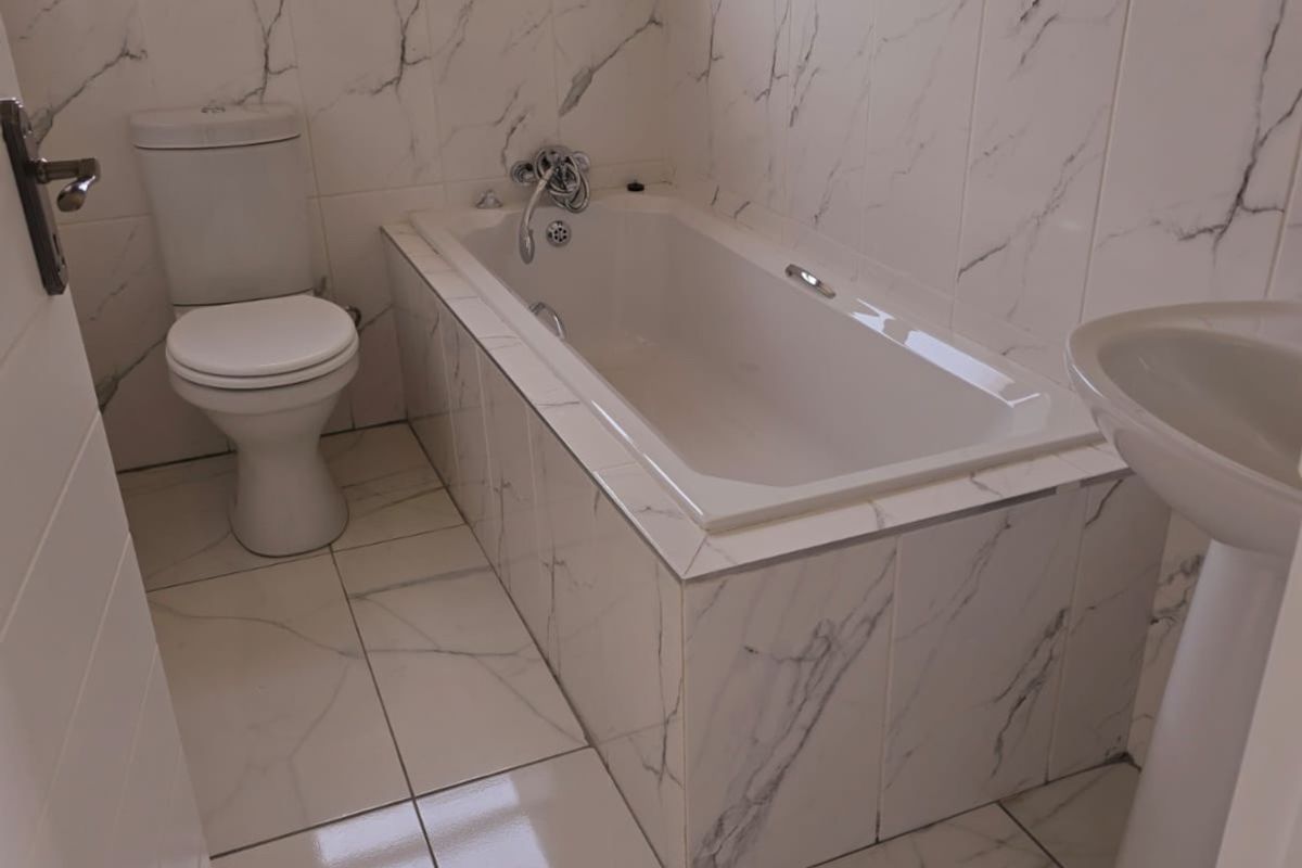 A clean, modern bathroom featuring marble-look tiled walls and floors, a built-in bathtub, a pedestal basin, and a standard toilet.