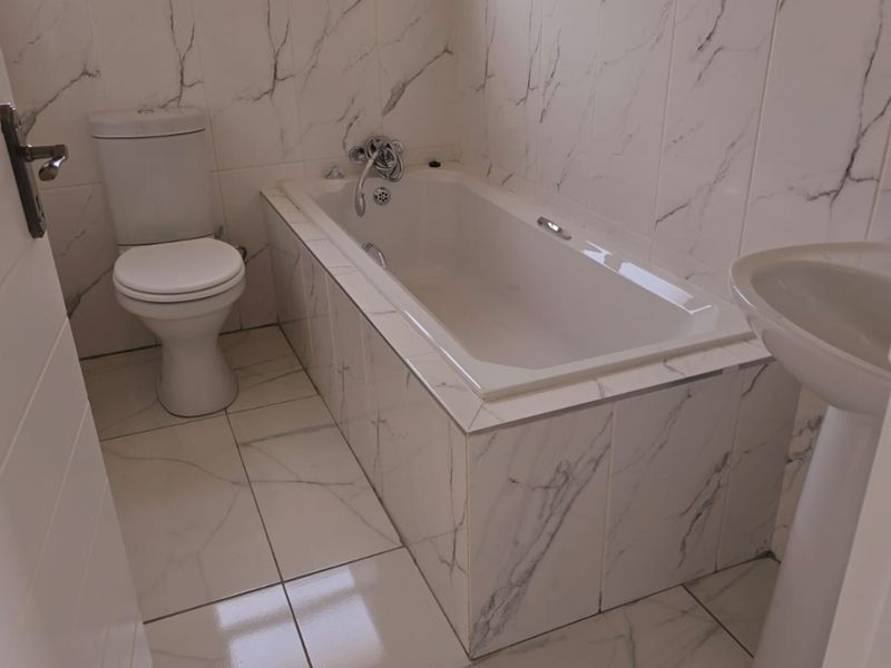 A clean, modern bathroom featuring marble-look tiled walls and floors, a built-in bathtub, a pedestal basin, and a standard toilet.