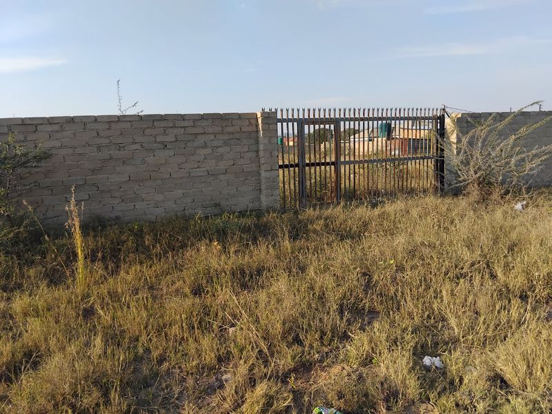 A view of a vacant residential plot featuring a brick boundary wall and a metal security gate with visible signs of weathering.