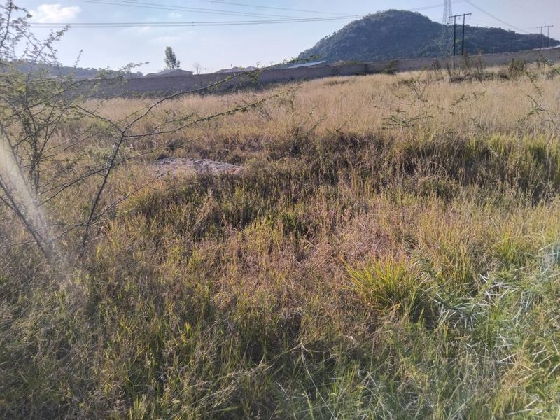 A view of a vacant residential plot featuring natural grass coverage, a perimeter wall, and distant mountain views.