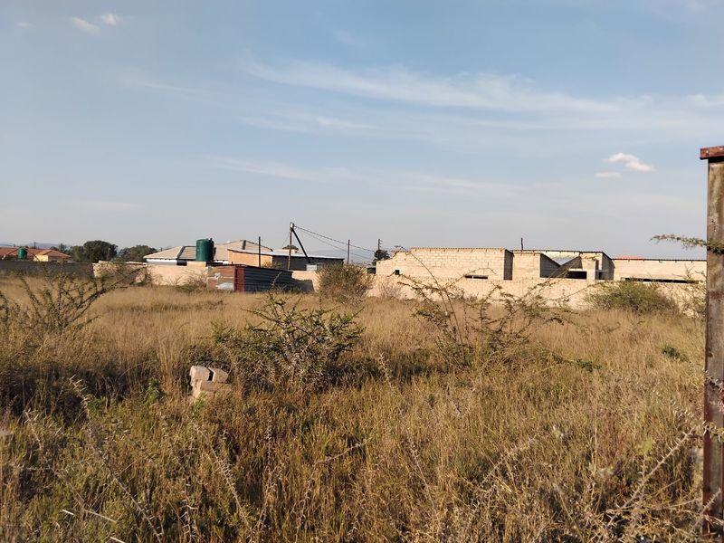 A view of a vacant residential plot featuring dry vegetation, a perimeter wall, and a water tank in the background.