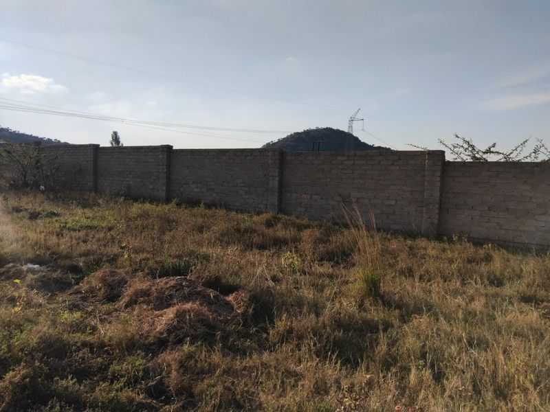 A vacant residential plot featuring a perimeter brick wall and natural, overgrown vegetation with distant mountain views.