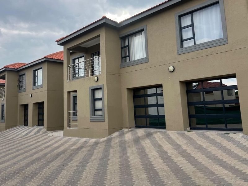 Front elevation of a modern townhouse complex featuring a paved driveway, double garage, and upper-level balcony with metal railings.